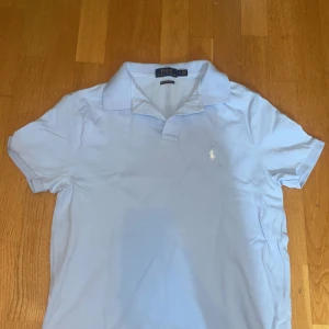 Ljusblå pikétröja från Polo Ralph Lauren - Snygg ljusblå pikétröja från Polo Ralph Lauren i bomull. Klassisk krage, korta ärmar och vit broderad logga på bröstet. Modellen är custom slim fit och passar perfekt till en clean och fräsch stil.