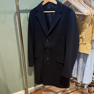 Mörkblå rock från Oscar Jacobson - Stilren mörkblå rock från Oscar Jacobson med klassisk krage och tre knappar framtill. Jackan har två fickor med lock och är helfodrad för extra komfort. Perfekt för dig som vill ha en tidlös och snygg look under kallare dagar. Storlek M/48 väldigt bra skick ny pris cirka 6000