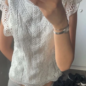 Vit spetsblus med volangdetaljer - Superfin vit blus i spets med snygga volangdetaljer vid axlarna och vackert mönstrad struktur.