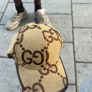 Gucci beige keps med GG-mönster - Snygg beige keps från Gucci med det klassiska GG-mönstret i brunt över hela kepsen. Kepsen har böjd skärm och bruna detaljer längs kanterna. Perfekt accessoar för att lyfta din streetwear-look.