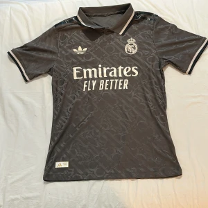 Real Madrid Vini Jr. matchtröja Adidas - Snygg mörkgrå Real Madrid fotbollströja med Vini Jr. och nummer 7 på ryggen. Tröjan har korta ärmar, krage och diskret mönster i tyget. Adidas-logga och klubbmärke på bröstet, samt Emirates Fly Better-tryck. Perfekt för dig som älskar fotboll och Real.