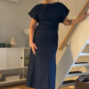 Mörkblå långklänning i linne - Snygg mörkblå långklänning i linne med korta ärmar och rund halsringning. Klänningen har en rak passform med markerad midja och en diskret slits på sidan. Perfekt för dig som gillar minimalistisk och stilren design.
