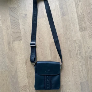 Svart axelväska från Philipp Plein - Snygg svart axelväska från Philipp Plein med justerbart band där loggan är broderad. Väskan har lock med magnetstängning och diskreta detaljer framtill. Perfekt storlek för mobil, plånbok och andra essentials.