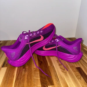 Nike | Men's Pegasus Plus Road Running Shoes - Vivid Purple - Nike Flyknit sneakers i en riktigt snygg lila färg med rosa detaljer och svart mesh. Skorna har en sportig vibe, platt sula och snörning. Perfekta för dig som vill sticka ut med färg på fötterna och gillar lätta, ventilerande material.
