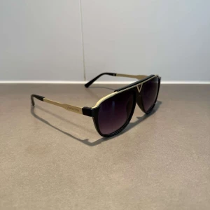 Louis Vuitton svarta solglasögon - Snygga solglasögon från Louis Vuitton med svart båge och gulddetaljer. Modellen har en markerad, rak ovansida och mörka tonade glas. Sidorna är prydda med Louis Vuitton-loggan i guld. Perfekt accessoar för att lyfta din stil.
