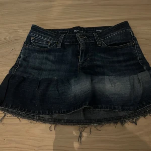 Levi's jeanskjol med rå kant - Mörkblå jeanskjol från Levi's med klassiska bakfickor och rå, fransig nederkant. Modellen är kort och har en lätt utställd volang nedtill. Perfekt för dig som gillar en avslappnad och cool denimstil.