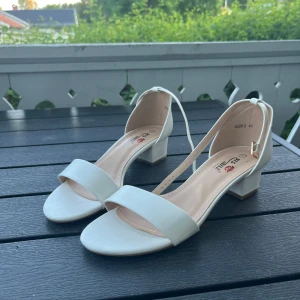 Vita sandaler med låg klack - Snygga vita sandaler från Be Mine med öppen tå och bred rem framtill. Skorna har en bekväm låg klack och justerbart spänne runt ankeln. Perfekta för sommarens alla tillfällen och lätta att matcha med olika outfits.