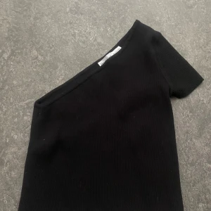 Svart oneshoulder topp från Bershka - Svart ribbad topp från Bershka med oneshoulder-design och kort ärm. Snygg och enkel modell som sitter tight och har en stilren look. Perfekt att styla med jeans eller kjol för en trendig vibe. Oanvänd, då den aldrig kommit till användning.