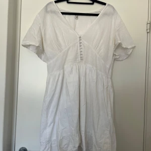 Vit kortklänning i linne med knappar - Superfin vit kortklänning i linne med v-ringning, korta ärmar och dekorativa tygklädda knappar framtill. Dragkedja på vänster sida. Viss missfärgning under armar efter deo, inget som syns vid användning.