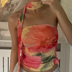 Blommig bandeau tubtopp med scarf - Superfin bandeau tubtopp med blommigt mönster i rosa, rött, grönt och gult. Toppen har en scarf som knyts runt halsen för en unik look. Sitter tight och är perfekt för varma dagar eller när du vill sticka ut lite extra.