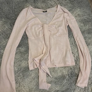 Ljusrosa omlottopp från Gina Tricot - Supersöt ljusrosa omlottopp från Gina Tricot med långa ärmar och knyt framtill. Tunt och mjukt material som känns skönt mot huden. Perfekt för dig som gillar en feminin och trendig look med v-ringning och snygg passform.