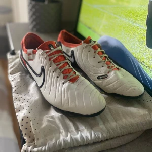 Nike Tiempo fotbollsskor vit/orange - Säljer ett par Nike Tiempo fotbollsskor i vitt med orange och svarta detaljer. Skorna har snörning, svart Swoosh-logga och textilpartier i orange. Ovandelen är i skinn och flyknit-material för extra komfort och passform. Perfekta för dig som vill ha en otroligt bekväm känsla på planen. Skick 9/10.
