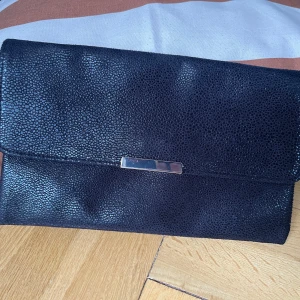 Svart kuvertväska med kedja - Stilren svart kuvertväska med strukturmönstrad yta och silverfärgad kedja som axelrem. Väskan har magnetlås och en liten metallplatta framtill. Perfekt storlek för det viktigaste och enkel att bära över axeln.