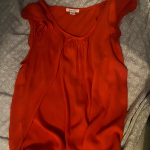 Röd ärmlös blus från Amisu - Snygg röd blus från Amisu med volangdetaljer vid axlarna och lätt rynk vid halsringningen. Blusen är ärmlös och har en mjuk, flowy känsla som gör den perfekt för varma dagar. Enkel att matcha med både jeans och kjol.