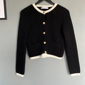 Svart kofta med guldknappar från Zara - Snygg svart kofta från Zara med vita kanter och dekorativa guldknappar framtill. Koftan är stickad, har två fickor och långärmad passform. Perfekt för lite kallare kvällar.