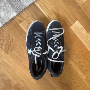 Svarta Converse One Star sneakers - Svarta Converse One Star sneakers i mocka med vit stjärna på sidan och vita snören. Klassisk låg modell med vit sula och rund tå. Perfekta för dig som gillar streetstyle och vill ha en ikonisk sneaker med cool retrovibe.