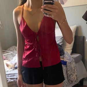 Röd satinlinnetopp med snörning - Snygg röd linnetopp i satin med smala axelband och djup v-ringning. Toppen har snörning framtill och är ärmlös med öppen rygg. 