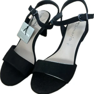 Svarta klackeskor från Tamaris - Säljer ett par svarta klackeskor från Tamaris med medelhög klack och öppen tå. Skorna har en enkel rem över tårna och justerbar rem runt vristen med spänne. Tillverkade i syntetmaterial och märkta som veganska. Aldrig använda endast testade storlek 37☺️nypris 699kr