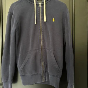 Marinblå hoodie från Polo Ralph Lauren - Säljer en marinblå hoodie från Polo Ralph Lauren med dragkedja och vit snörning i huvan. Klassisk gul broderad logga på bröstet och två fickor framtill. Skön bomullskvalitet och perfekt för chill dagar.