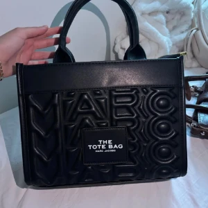 Svart handväska Marc Jacobs Tote - Svart handväska från Marc Jacobs med präglad logotyp över hela väskan och tydlig patch framtill med texten 'The Tote Bag'. Väskan har två stadiga handtag och är tillverkad i syntetskinn. 💕