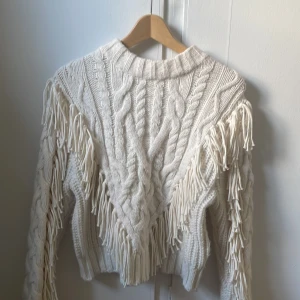 Beige stickad tröja med fransar - Säljer en beige stickad tröja med grovt kabelstickat mönster och coola fransdetaljer längs ärmar och fram. Tröjan har lång ärm och en lite boxig passform, perfekt till ett par jeans shorts!