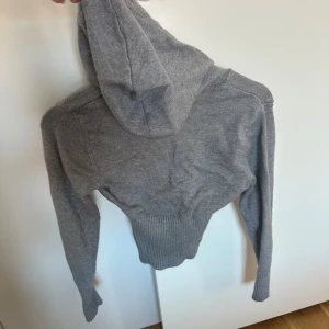 Grå stickad hoodie från Gina Tricot - Grå stickad hoodie från Gina Tricot i storlek XS. Tröjan har dragkedja framtill, snörning i huvan och ribbade muddar samt midja. Perfekt för dig som gillar en enkel och stilren look med en mysig känsla.