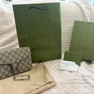 Gucci Dionysus GG Supreme kuvertväska - Snygg kuvertväska från Gucci i beige och brunt GG Supreme-mönster. Väskan har en ikonisk silverfärgad spänne med ormdetaljer och kedjerem. Insidan har flera kortfack och dragkedjefack. Perfekt för dig som vill ha en lyxig och stilren accessoar. Väskan är iprincip oanvänd och i ett otroligt bra skick. Påse, låda, dustbag samt kvitto tillkommer. Orginalpris : 15.200kr men säljer den för 10.500 men pris kan även diskuteras.