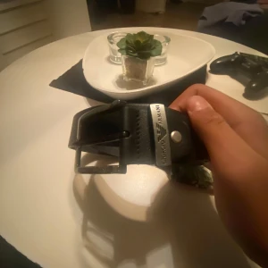 Svart skärp från Giorgio Armani - Snyggt svart bälte från Giorgio Armani i äkta skinn med robust silverfärgat spänne där loggan är ingraverad. Bältet har en klassisk design med bred rem och flera hål för justering. Perfekt accessoar för att lyfta din outfit.