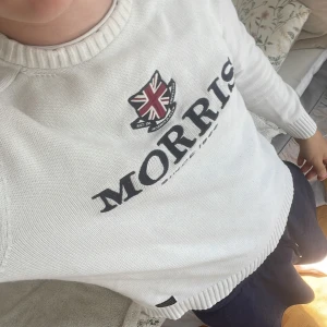 Vit stickad tröja från Morris - Vit stickad tröja från Morris med broderad logga och brittisk flagga på bröstet. Tröjan har rund halsringning, långa ärmar och ribbade muddar. Snygg och klassisk stil med tydlig text 'MORRIS SINCE 1968' framtill.