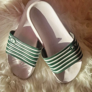 KV tofflor i grönt och vitt - Snygga slides från KV med gröna och vita ränder över foten. Skorna har en platt sula och är tillverkade i syntetmaterial. Perfekta för sommardagar eller att chilla hemma. Klassisk design med öppen tå och bekväm passform. Kommer från anstalten Hinseberg