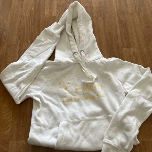 Vit hoodie Verbier Mont Fort - Snygg vit hoodie med guldig text 'Verbier Mont Fort Switzerland' på bröstet. Tröjan har huva med snörning och en stor magficka. Perfekt för dig som gillar en clean och stilren look med en touch av lyx.