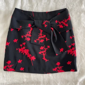 Svart minikjol med röd blommig print Ivyrevel - Supersnygg svart kortkjol från Ivyrevel med rött blommönster och stor rosett framtill i midjan. Kjolen har en figurnära passform och dragkedja i sidan. Längd: 41cm, Midja: 34cm