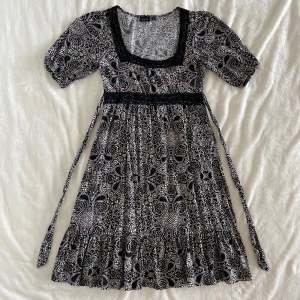 Mönstrad klänning Lindex S - Supersöt svart/brun/vit/beige knälång klänning från Lindex med unikt mönster, markerad midja med knytband och spetsdetaljer vid halsringningen. Puffiga korta ärmar och flowig kjol. Längd: 97cm, Midja: 37cm