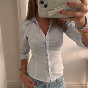 Randig blåvit skjorta med trekvartsärm - Snygg figursydd skjorta från H&M i blåvitt randigt mönster. Skjortan har trekvartsärmar, klassisk krage och knäppning framtill. 