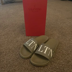 Valentino Garavani slides i khaki - Snygga slides från Valentino Garavani i khaki-grön färg med vit VLTN-logga över remmen. Tillverkade i syntetmaterial med platt sula och öppen tå. Perfekta för sommaren och riktigt trendiga att bära till shorts eller jeans.