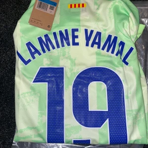 FC Barcelona bortatröja 23/24 Nike - FC Barcelona bortatröja säsong 23/24 med tryck Lamine Yamal 19 eller Raphinha 11. Tröjan är ljusgrön med abstrakt mönster, blå detaljer och klubbmärke på bröstet. Tillverkad i lätt och ventilerande material, kortärmad och med sponsorloggor från Spotify och UNHCR.