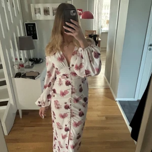 Baloon sleeve dress från gina  - Säljer en elegant vit långklänning med rosa blommigt mönster. Klänningen har långa ärmar och en v-ringad design som ger en stilren look. Perfekt för speciella tillfällen. Helt slutsåld på Gina 