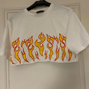 Vit croppad t-shirt med eldtryck från Shein - Säljer en cool vit croppad t-shirt från Shein med ett eldtryck i gult och rött längs nederkanten. Perfekt för en trendig look. T-shirten har korta ärmar och rund halsringning.