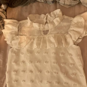 Vit blus med volangdetaljer - Säljer en söt vit blus med volangdetaljer och prickigt mönster. Blusen är kortärmad och har en transparent övre del med en hög krage. Perfekt för en stilren och romantisk look.