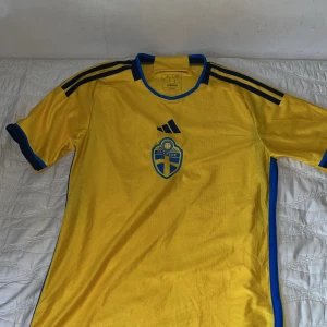 Gul fotbollströja från Adidas - Säljer en gul fotbollströja från Adidas med blå detaljer och korta ärmar. Tröjan har ett emblem på bröstet och tre blå ränder på axlarna. Perfekt för fotbollsfans!