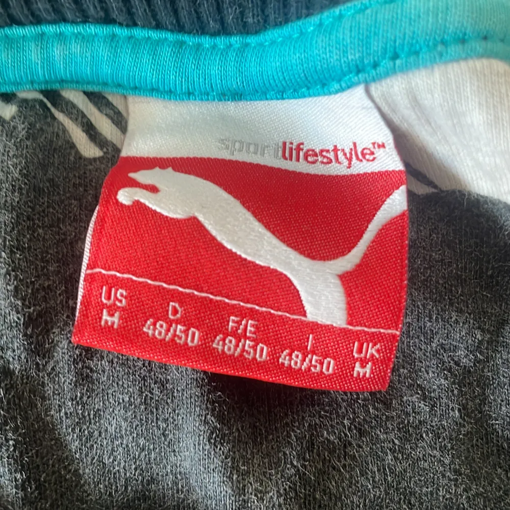 Säljer en mörkblå långärmad tröja från Puma med ett stort logotryck i turkos och vitt på framsidan. Tröjan har en rund halsringning och ribbade muddar vid ärmslut och nederkant. Perfekt för en sportig look ✅. Neuletakit & Villapaidat.