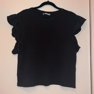 Svart topp med volangärmar från Zara - Säljer en stilren svart topp från Zara med korta volangärmar. Toppen har en rund halsringning och är i storlek S