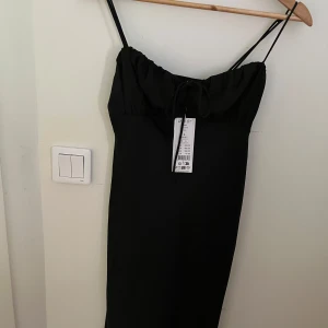 Svart klänning med smala axelbands  - Elegant svart klänning med smala axelband och knytning framtill. Perfekt för en stilren look. Klänningen har en lång design som geri en sofistikerad känsla.