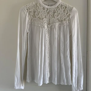 Vit blus med spetsdetaljer - Elegant vit blus med vackra spetsdetaljer på axlar och bröst. Blusen har långa ärmar och en knapp i nacken för en stilren look. (Katt och kanin finns i hemmet)