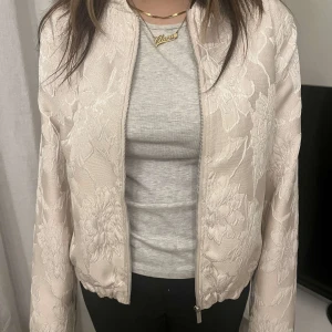 Beige blommig bomberjacka - Säljer en snygg beige bomberjacka med blommigt mönster. Jackan har dragkedja framtill och långa ärmar. Den är storlek 40 men passar bättre som 36.