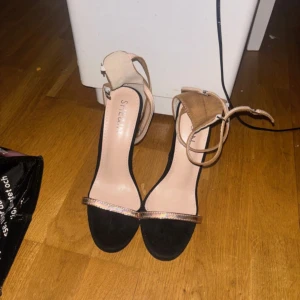 Svarta och beige klackskor från NLY - Snygga klackskor från NLY med svart och beige design. Skorna har en elegant rem över tårna och en justerbar rem runt ankeln. Perfekta för en kväll ute eller en speciell tillställning.