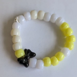 Färgglatt pärlarmband med rosett - Charmigt armband med vita och gula pärlor samt en svart rosett. Perfekt för att lägga till en lekfull touch till din outfit. Passar barn och ungdomar 