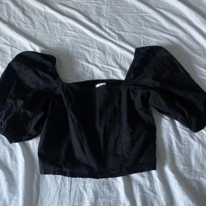 Svart blus från H&M - Snygg svart blus från H&M med puffiga ärmar och broderade detaljer. Toppen har en smockad rygg och är offshoulder, vilket ger en trendig look. Perfekt för att lyfta vilken outfit som helst!
