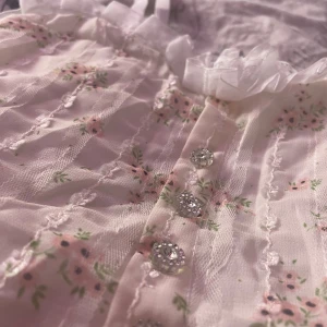 Blommig topp med spetsdetaljer - Säljer en romantisk topp med blommigt mönster i rosa och grönt. Toppen har vackra spetsdetaljer och glittrande knappar framtill. Perfekt för en söt och feminin look. Köpt i Thailand och är knappt använd