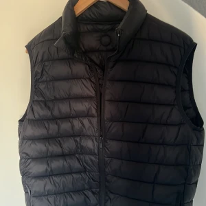 Svart väst från kronstadt  - Snygg svart dunväst med dragkedja framtill och hög krage. Perfekt för kyligare dagar och enkel att matcha med andra plagg. Västen har en quiltad design som ger en stilren look. I storlek M och har inte använts.